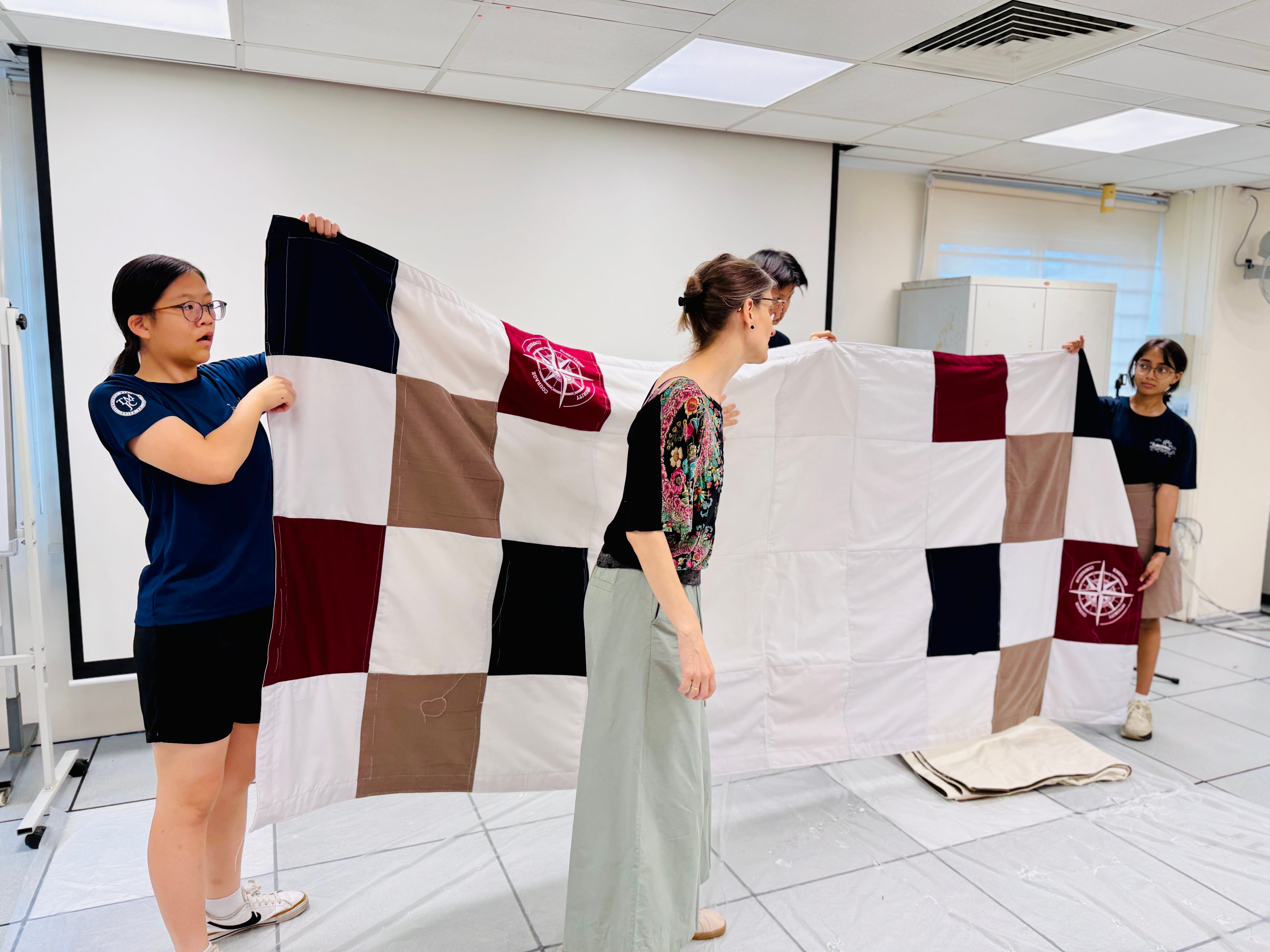 Three people hold up a large patchwork quilt-style banner made of coloured fabric squares in white, navy, maroon, and tan, featuring a compass rose emblem. They are standing indoors in what appears to be a classroom or workroom.